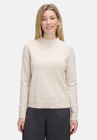 Glunderende vrouw met stijlblond haar, gekleed in een lichtbeige lange mouwtrui en donkere losse broek tegen een eentonige achtergrond.