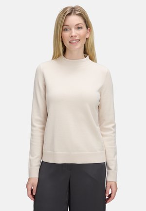Lächelnde Frau mit glattem, blondem Haar, die einen hellbeigen, langärmligen Pullover und dunkle, lockere Hosen trägt, vor einem schlichten Hintergrund.