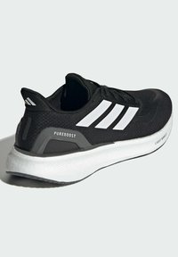 Chaussure de sport noire avec des rayures blanches, tige en maille, semelle rembourrée et accents gris. Présente des étiquettes "PUREBOOST" et "LIGHTBOOST".
