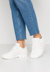 Vita nät sneakers i vitt med rosa rand detaljer, som har en texturerad ovandel och en vit sula. Bärs tillsammans med blå jeans med uppvikta ben.