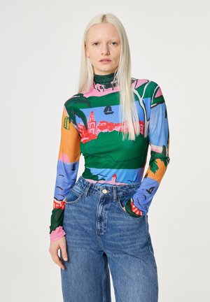 Persoon die een kleurrijk, met een landschap bedrukt, top met lange mouwen en een hoge hals draagt, ingestopt in een high-waisted blauwe spijkerbroek, hand in de zak, neutrale achtergrond.