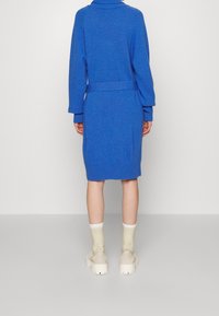 Vestito blu a maglia con colletto alto e vita stretta da una cintura. Presenta polsini e orlo a costine, design fino al ginocchio, abbinato a calzini crema alla caviglia e scarpe dal tacco massiccio.