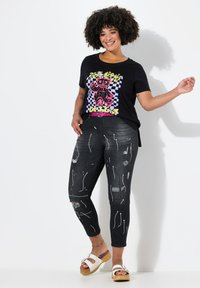 Camiseta negra con un gráfico colorido que presenta un oso sobre un fondo de tablero de ajedrez, combinado con jeans negros desgastados con un diseño de salpicaduras blancas.