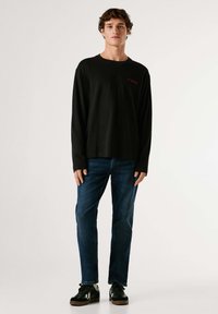 Črna srajca z dolgimi rokavi z rdečim napisom, kombinirana s temno modrimi kavbojkami in črnimi supergami z belimi poudarki. Model stoji na enostavni podlagi.