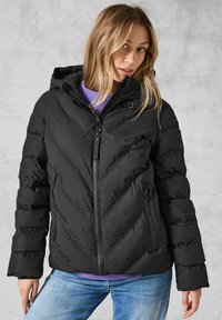 Giacca a vento nera imbottita con zip davanti, motivo a zigzag e cappuccio. Presenta tasche laterali e tessuto testurizzato. Indossata sopra un maglione viola.