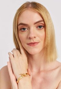 Zlatý kovový náramok s uzlovým akcentom, nosený na ruke so svetlou pokožkou, skombinovaný s neutrálnym líčením na jednotnom pozadí.