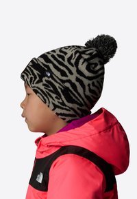 Czarny i szary dzianinowy czapka w pasy zebry z pomponem na czubku, noszona przez dziecko w jasnoróżowej kurtce z czarnymi akcentami.