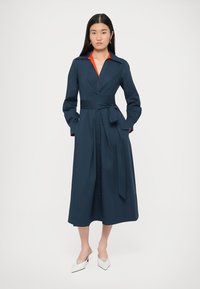 Marineblauwe midi-jurk met een getailleerde taille, lange mouwen, rode kraag en zijstrik. Voorzien van een knoopsluiting aan de voorkant en een uitlopende rok. Witte schoenen met hak.