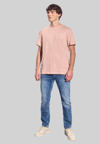 Světle růžové bavlněné tričko s kulatým výstřihem a krátkými rukávy, spárováno s modrými džíny a béžovými teniskami. Model stojí čelem.