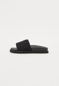 Sandale negre cu talpă plată din cauciuc, având o curea din material țesut și un design minimalist. Textură netedă cu detalii căptușite pe curea.