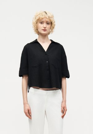 Γυναίκα με κοντά κυματιστά ξανθά μαλλιά που φοράει χαλαρό μαύρο πουκάμισο με κοντά μανίκια και άσπρο παντελόνι, με φόντο ένα απλό σκηνικό.