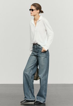 Woman wearing black sunglasses, white long-sleeve shirt tucked into loose blue jeans, holding a bag, standing on gray floor with light gray background.