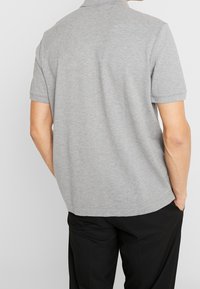 Polo shirt gris à manches courtes avec un col classique, texture lisse et ourlet droit. Porté avec un pantalon noir, montrant la vue arrière.