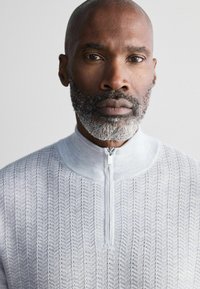 Hellgrauer Reißverschluss-Pullover mit einem strukturierten Zickzackmuster, weichem Strickmaterial, hohem Kragen und subtilen Glanzelementen.