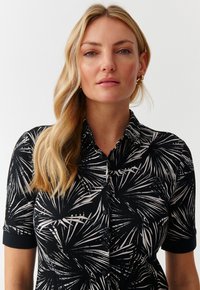 Camicia nera con motivo di foglie di palma bianche, maniche corte e chiusura con bottoni. Presenta un colletto classico e una texture di tessuto liscia.