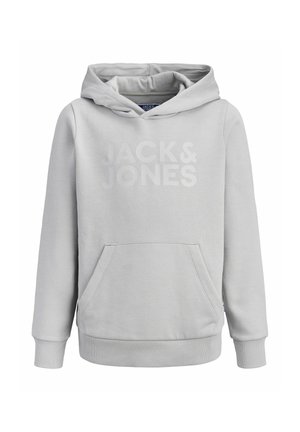 Mladić stoji, nosi svijetlo sivu majicu s kapuljačom, crne traperice, crno-bijele tenisice i crnu kapu s logom "Jack & Jones".
