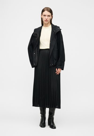 Femme aux cheveux bruns lisses portant un chemisier crème, une veste à capuche noire, une jupe midi plissée noire et des bottes à talons noires, debout.