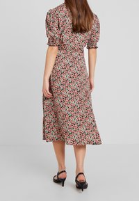 Žena v midi šatech s růžovým květinovým vzorem, s balónovými rukávy a černými sandály na podpatku, stojící na jednobarevném šedém pozadí.