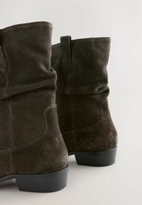 Bottines en daim marron foncé avec un design loose, des bouts ronds et des talons bas et épais noirs. Présente des coutures visibles et des languettes de tirage.