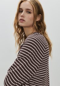 Ung kvinde med bølget lyst brunt hår iført en brun og hvidstribet langærmet trøje, der ser let over sin skulder.