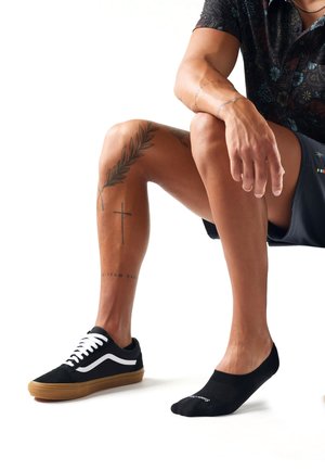 Homme assis, un pied portant une basket noire à lacets blancs et l'autre pied avec une chaussette noire invisible, tatouages visibles sur le bras et portant un short.