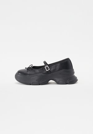 Chaussure Mary Jane noire avec semelle épaisse texturée, petit nœud sur le bout et bride à boucle réglable sur fond blanc.