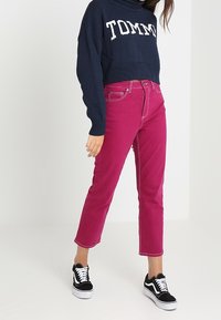 Marineblauer, gestrickter Cropped-Pullover mit "TOMMY" in weißem Text; fuchsiafarbene Jeans mit geradem Schnitt und Kontrastnähten; schwarze Sneakers.