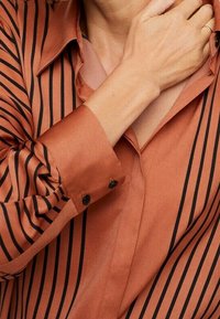 Persona che indossa una camicia arancione ruggine con strisce verticali nere, la mano poggiata delicatamente sul collo vicino alla clavicola.