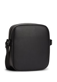 Μαύρη τσάντα ώμου από λείο συνθετικό υλικό. Τετράγωνο σχήμα με κλείσιμο με φερμουάρ, ρυθμιζόμενο ιμάντα και μινιμαλιστικό σχεδιασμό.