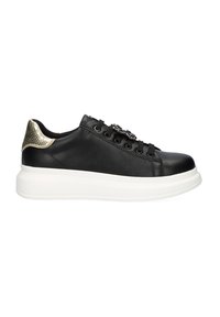 Zapatilla negra de cuero con suela blanca, con un acento en el talón de metal dorado y cordones decorativos. Textura suave, forma redondeada.