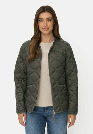 Kvinde med langt brunt hår iført en mørkegrøn quiltet jakke over en beige skjorte og blå jeans, stående mod en ensfarvet lys baggrund.