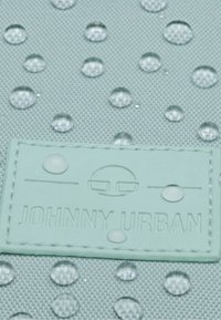Kapky vody na texturované světle modré tkanině s vyšitým štítkem s nápisem "Johnny Urban" a minimalistickým obličejovým logem.