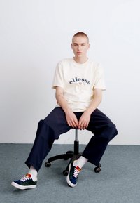 Camiseta beige con Logotipo azul, combinada con pantalones anchos oscuros. Zapatillas multicolores con acentos rojos, amarillos y verdes. Sentado en una silla.