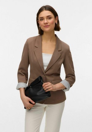 Žena s krátkymi hnedými vlasmi, ktorá nosí hnedé sako cez biely top a nohavice, drží v oboch rukách čiernu koženú kabelku.