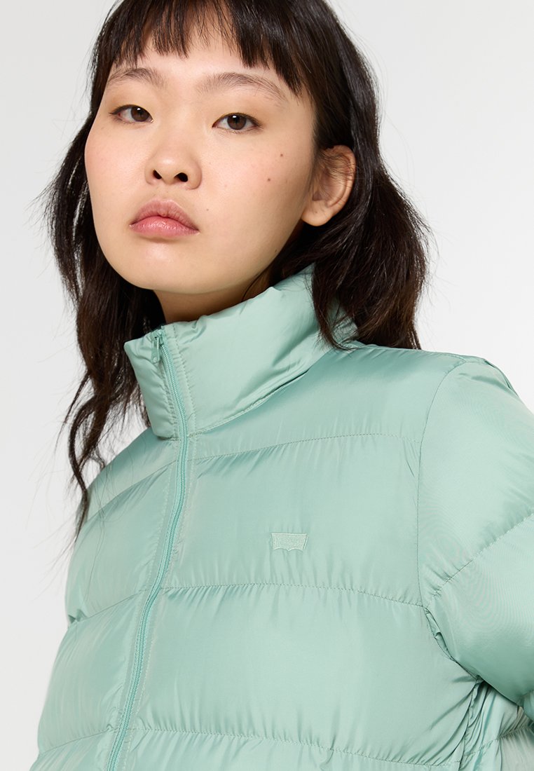 Chaqueta acolchada verde menta con un cuello alto, cierre de cremallera y una textura suave y brillante. Presenta un logo sutil en el pecho.
