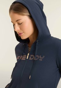 Giovane donna con felpa blu navy con cerniera e cappuccio alzato, occhi chiusi, rivolta a sinistra, con un'espressione calma e un trucco sobrio.