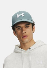 Gaiši zils cepures ar izliekto malu, kuru rotā balts Under Armour logo. Valkāta kopā ar pelēku kapuci, kas izceļ mīkstu tekstūru.