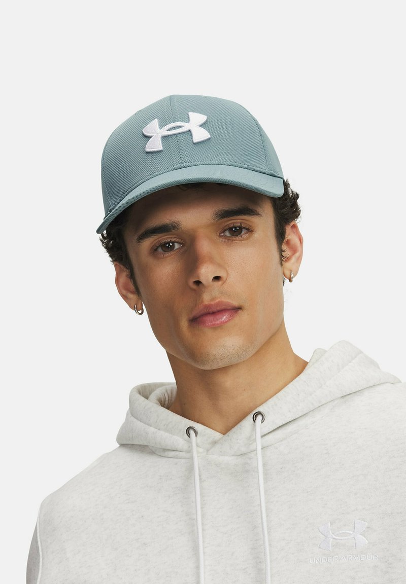 Gaiši zils cepures ar izliekto malu, kuru rotā balts Under Armour logo. Valkāta kopā ar pelēku kapuci, kas izceļ mīkstu tekstūru.