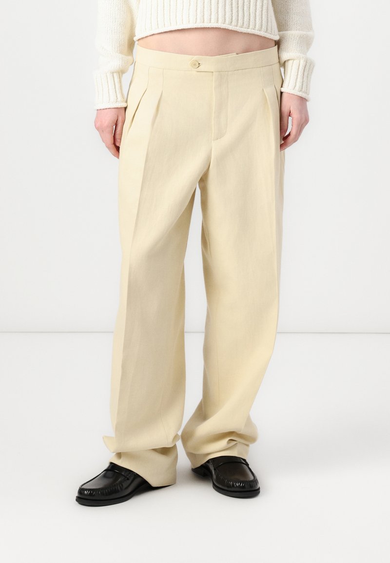 Crème kleurige geplooide broeken met een hoge taille, voorzien van een enkele knoopsluiting en rechte, losvallende pijpen. Draag ze met zwarte schoenen.