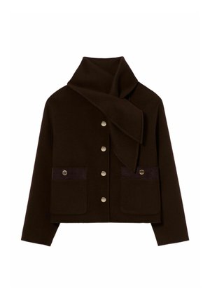 Persona de pie con chaqueta marrón oscuro con detalle de bufanda, pantalones anchos beige y zapatos marrones sobre un fondo claro y liso.