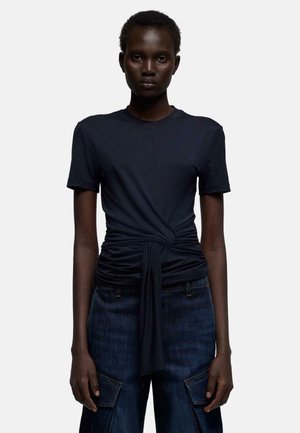 Woman wearing a dark navy short-sleeve shirt with a twisted knot detail at the waist and dark blue cargo jeans.