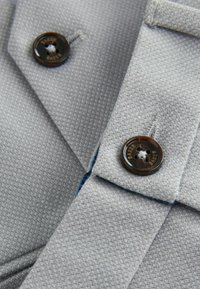 Tessuto grigio testurizzato con un motivo intrecciato, caratterizzato da un grande bottone marrone scuro con la scritta "BAKER" incisa. Cuciture visibili in blu.