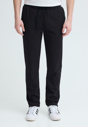 Mand stående iført sorte sweatpants med snor og hvide sneakers med sorte striber på en hvid baggrund.