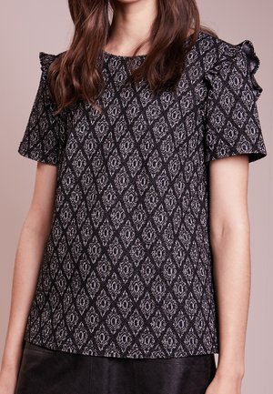 Femme portant un haut à manches courtes noir et blanc à motifs avec des détails volantés aux épaules sur un fond uni.