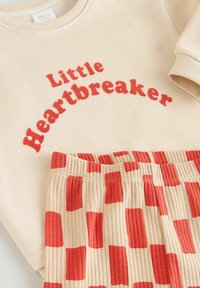 Beiger Pullover mit rotem Schriftzug "Little Heartbreaker", kombiniert mit beigen, gerippten Hosen mit abwechselnden roten rechteckigen Mustern.