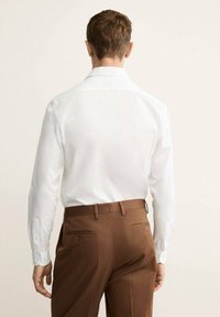 Chemise blanche à manches longues avec un col pointu, associée à un pantalon tailleur marron avec poches arrière et accents de boutons.