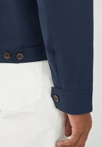 Marineblå jakke med en glat tekstur, der har to brune knapper ved mansjetterne. Hvid skjorte titter frem under jakken.