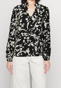 Svart och krämfärgad mönstrad blus med V-ringning, långa ärmar och avslappnad passform. Tyget verkar vara slätt med en lätt textur.