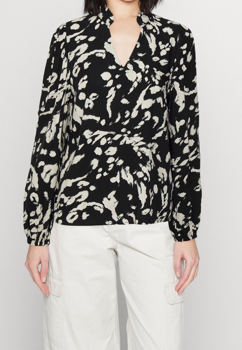 Svart och krämfärgad mönstrad blus med V-ringning, långa ärmar och avslappnad passform. Tyget verkar vara slätt med en lätt textur.