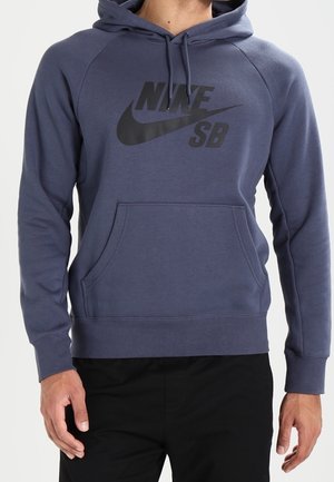 Mężczyzna w ciemnoniebieskiej bluzie z kapturem Nike SB z przednią kieszenią i czarnych spodniach, stoi na jednolitym jasnym tle.